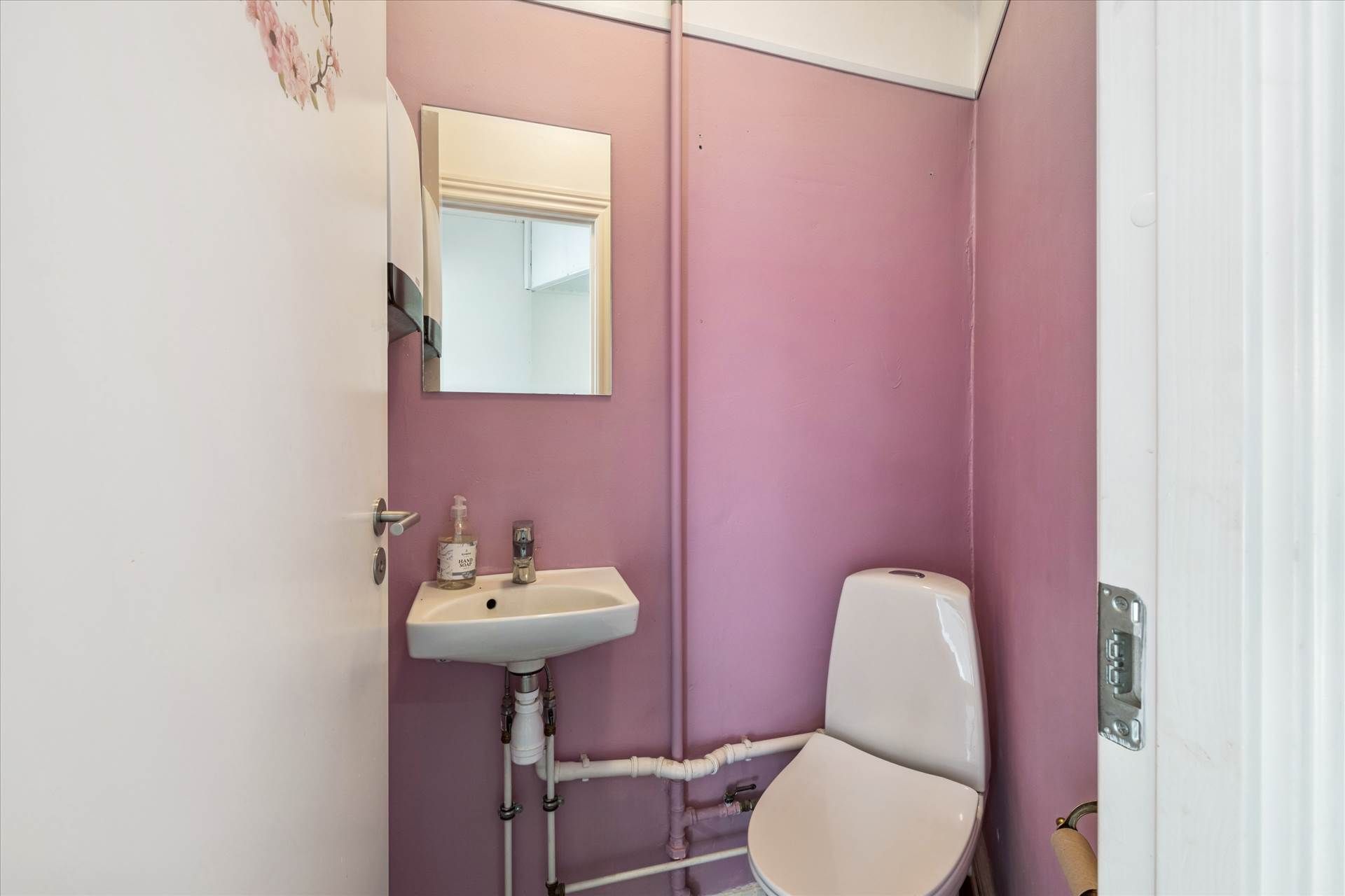 Toilet med hvidt toilet, hvidt vask og en spejl med en rørinstallation i baggrunden og en lyserød væg