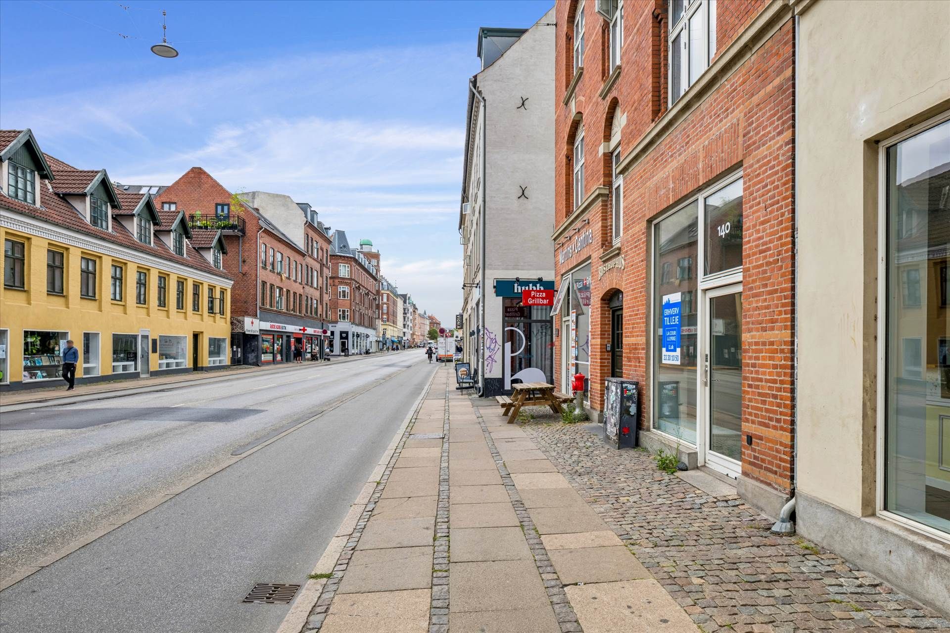 Butik med Pizza Grillbar skilt og rødt hydrant. Brede brolægget fortov og tom gadegade med spredte butikker. Gule og røde