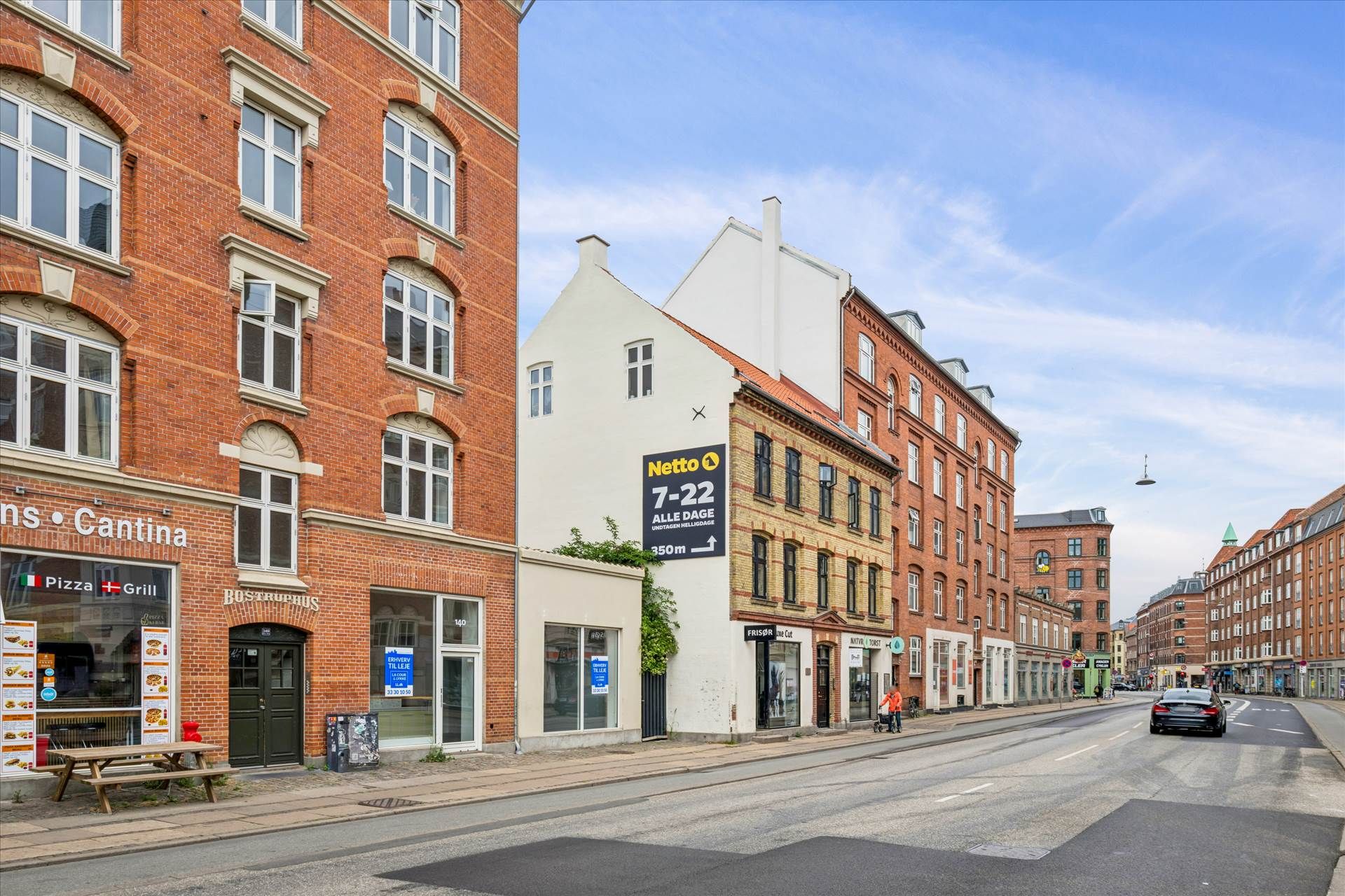 Stor bygning med restauranten Cantina og pizza-baren Bøstruphus. Butikker med reklame og skilte. En person går på fortovet.