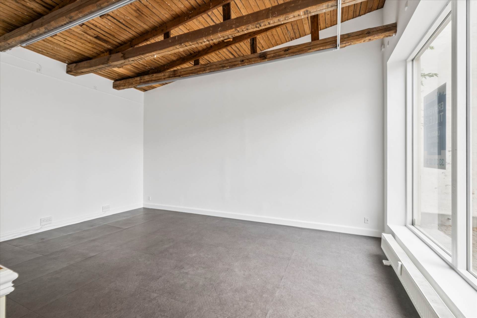 Indenfor er der et tomt rum med hvide vægge og en grå gulvbelægning, samt et træloft med metalbånd og en vinduesåbning med