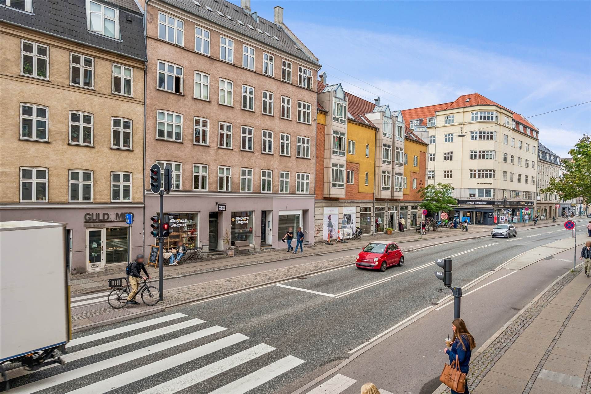 Bygning med flere butikker og caféer på Amagerbrogade. Mennesker går og cykler på gågaden. En bil og en lastbil kører på