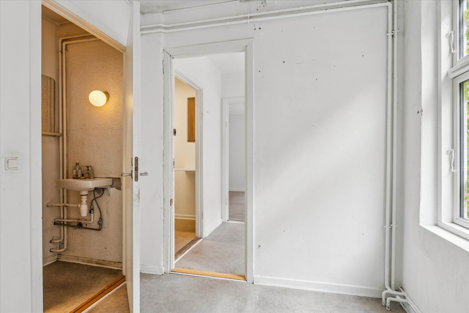 Et tomt værelse med åbne døre og en åben vindueskilt og et lille badeværelse med en hvid vask og en vandhane og et gulv med