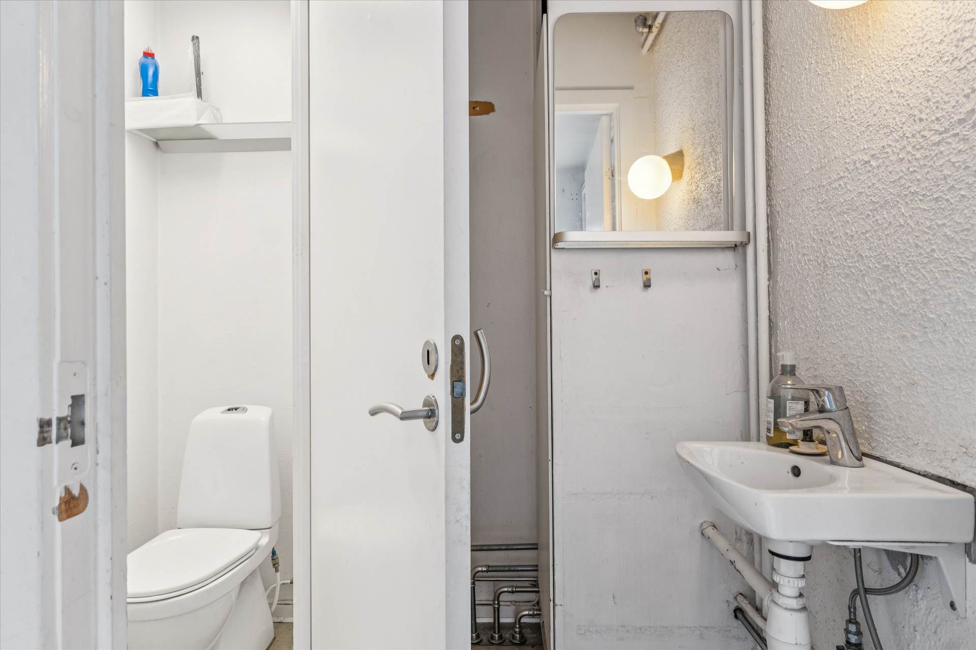 Et lille, rent og hvidt badeværelse med toilet, håndvask og spejl samt et toiletpapirhylde med en plastflaske ovenpå