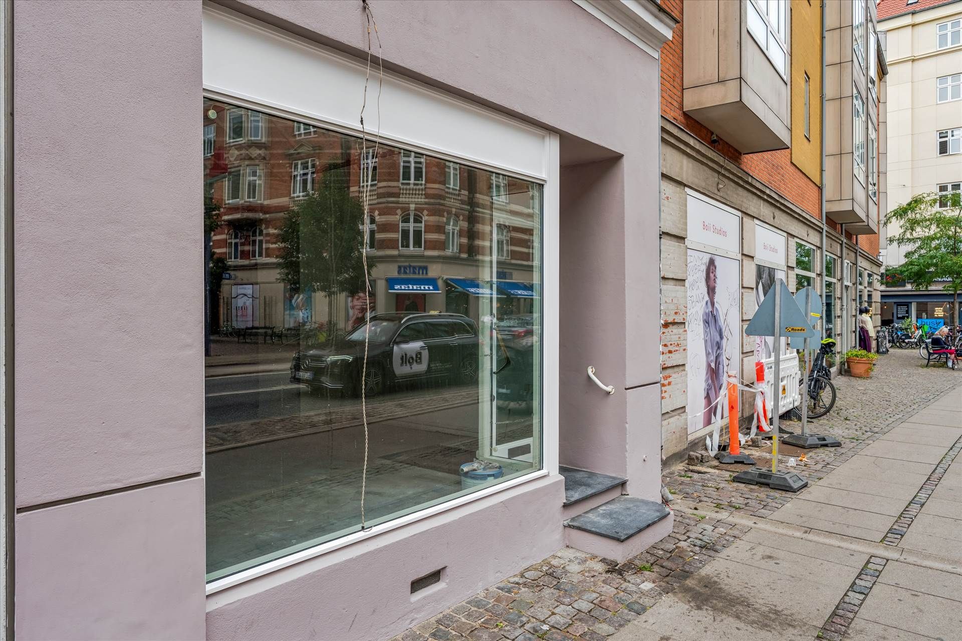 Lejlighed med grønne ruder og glasdør i bygning. Bagved er der en skrågang med skrald, og en cykel står parkeret.