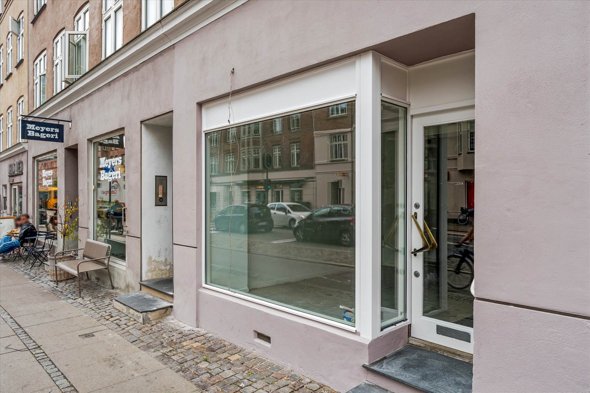 Bygningen med glasvinduer og hvide indramninger på Amagerbrogade 48, to personer sidder og nyder kaffe udenfor og en cykel