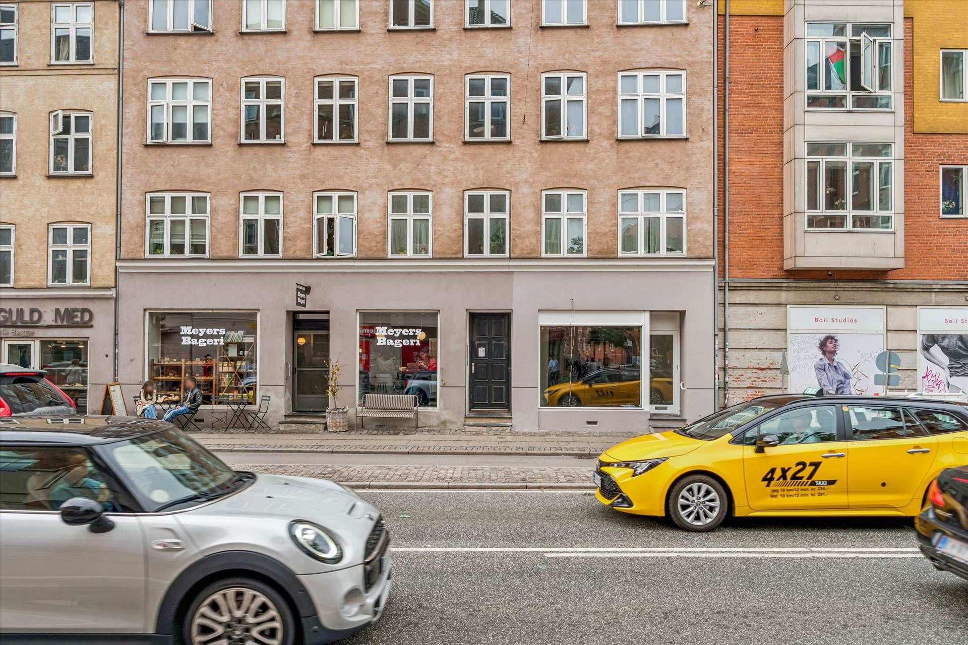 To biler kører på en gade med butikker og et gult taxibil med nummerplade 4x27