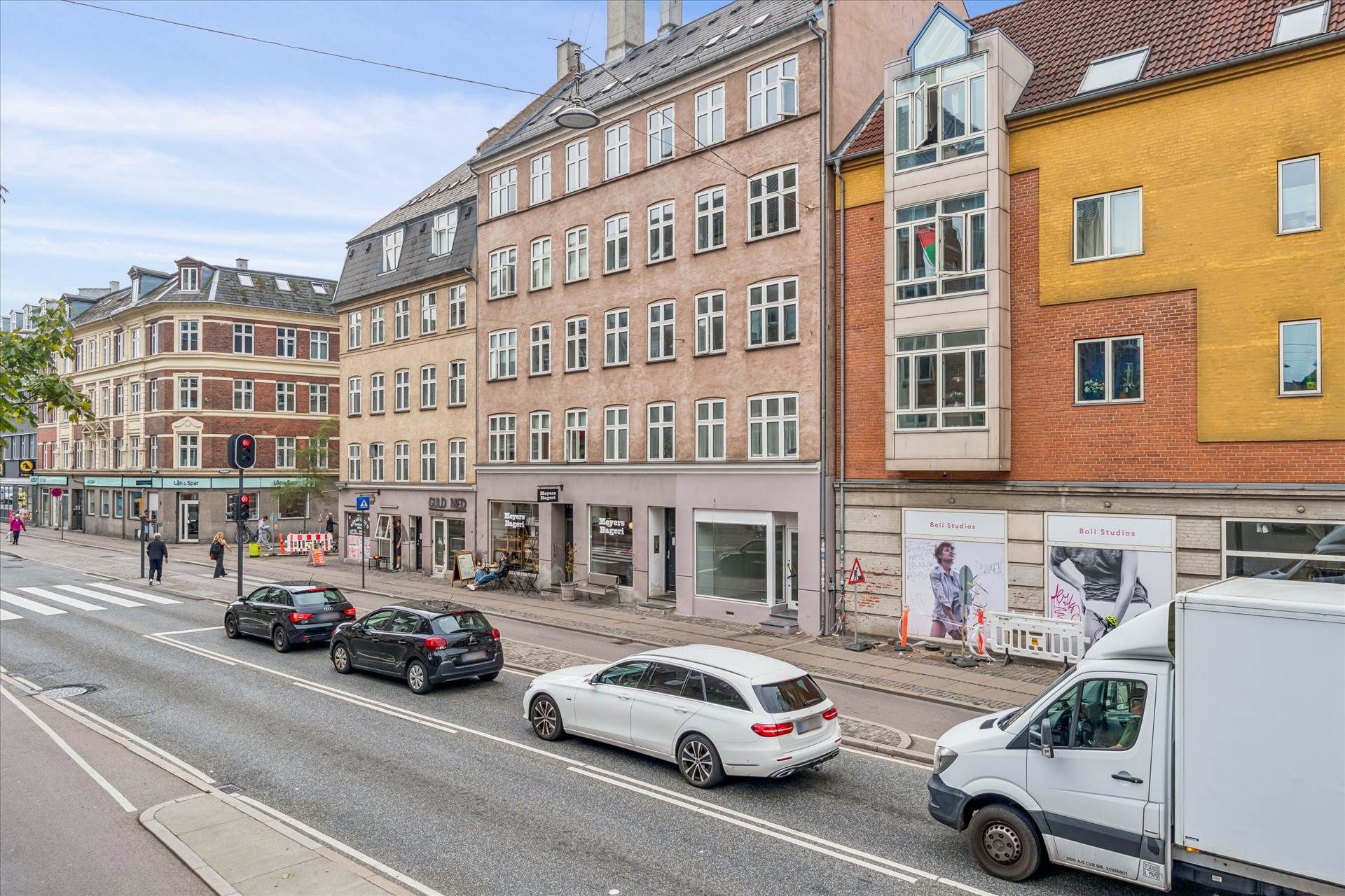 Flere biler kører langs en handelsgade, og der er en stor lastbil, der kører foran, samt forskellige bygninger langs vejen.