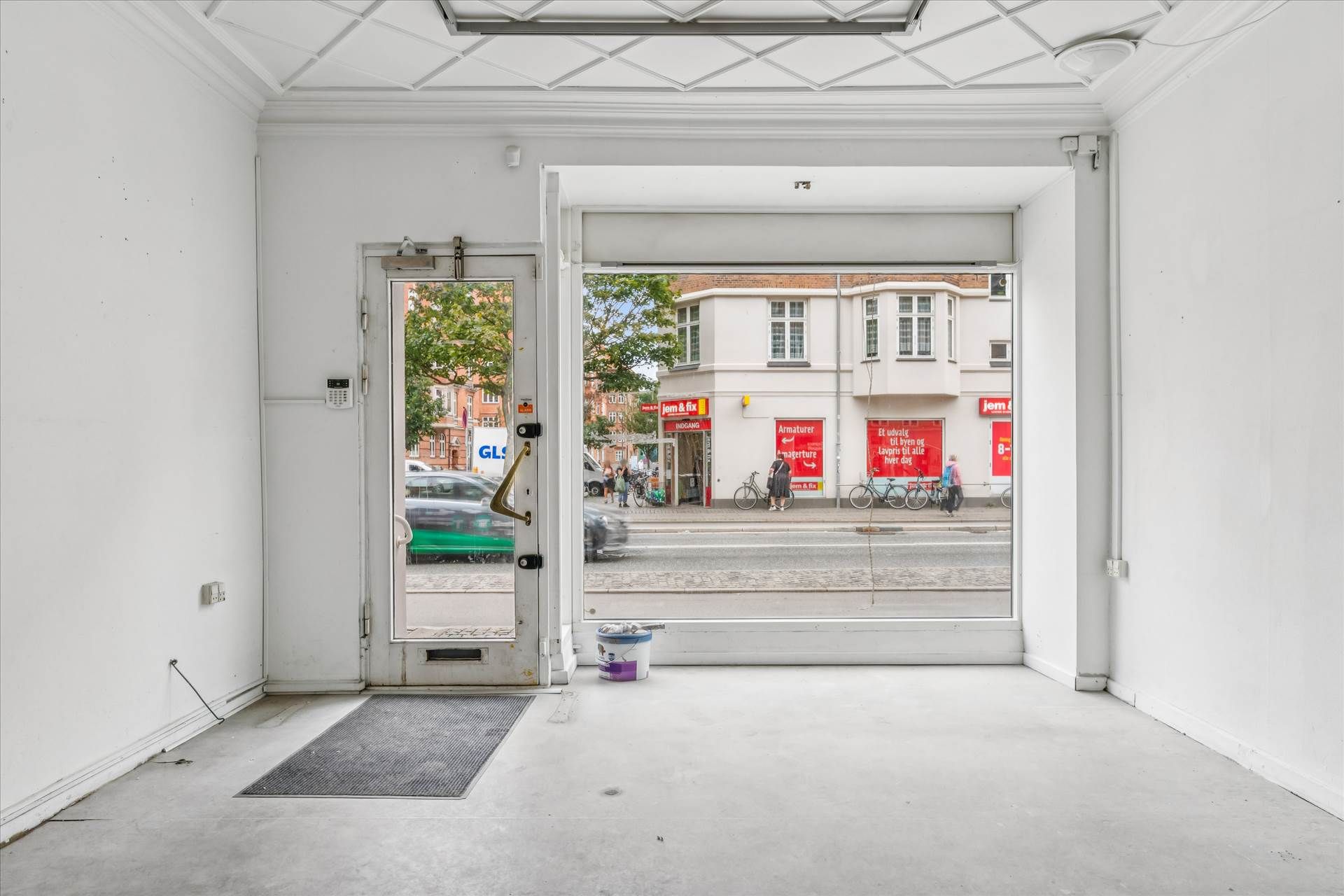 To store glasdøre med åbning mod gaden med butikker, hvor folk går forbi, og en bil kører lige forbi