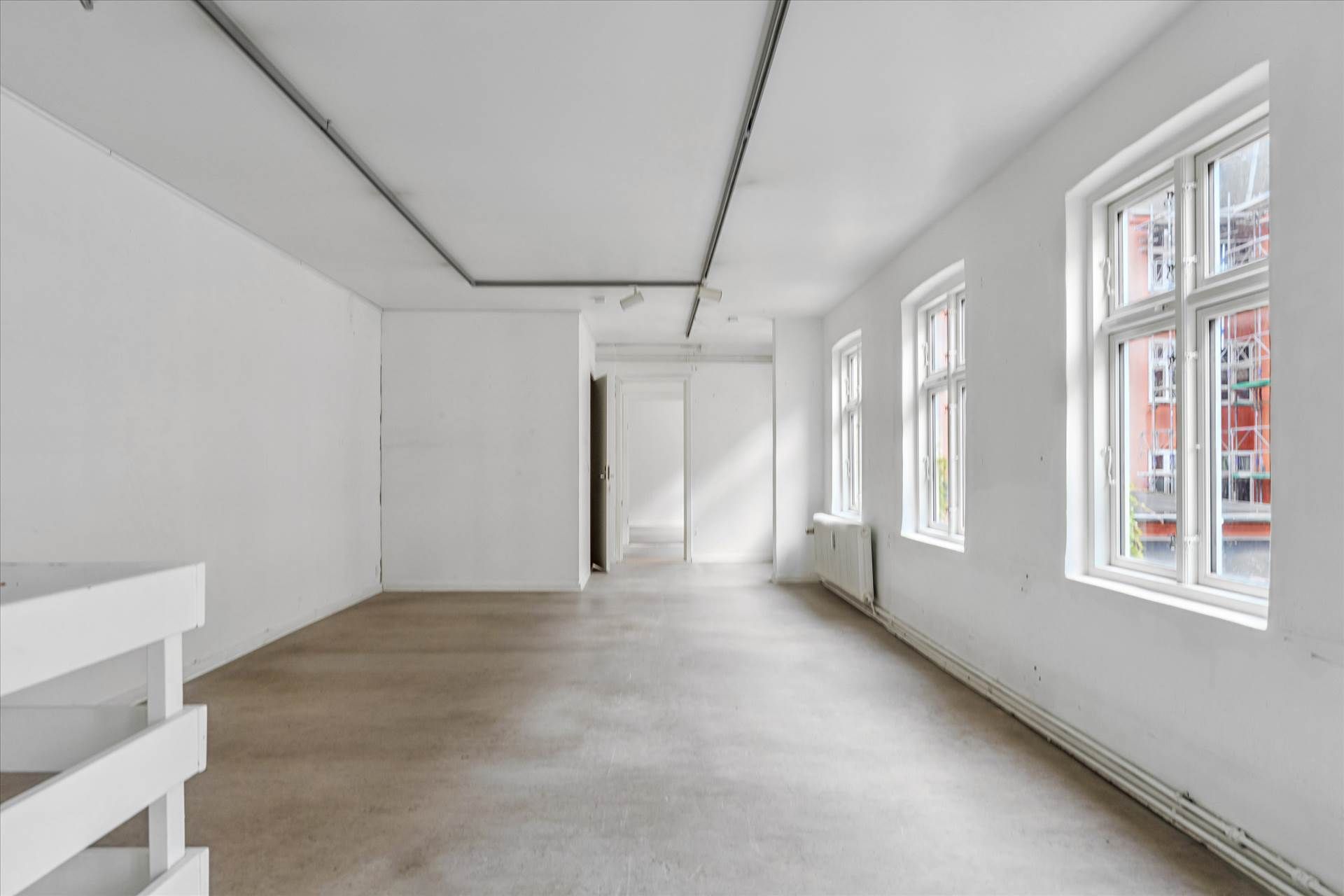 En stor, tom lejlighed med hvide vægge, lysegrå gulv og vinduer, samt åbne døre mod en gang.