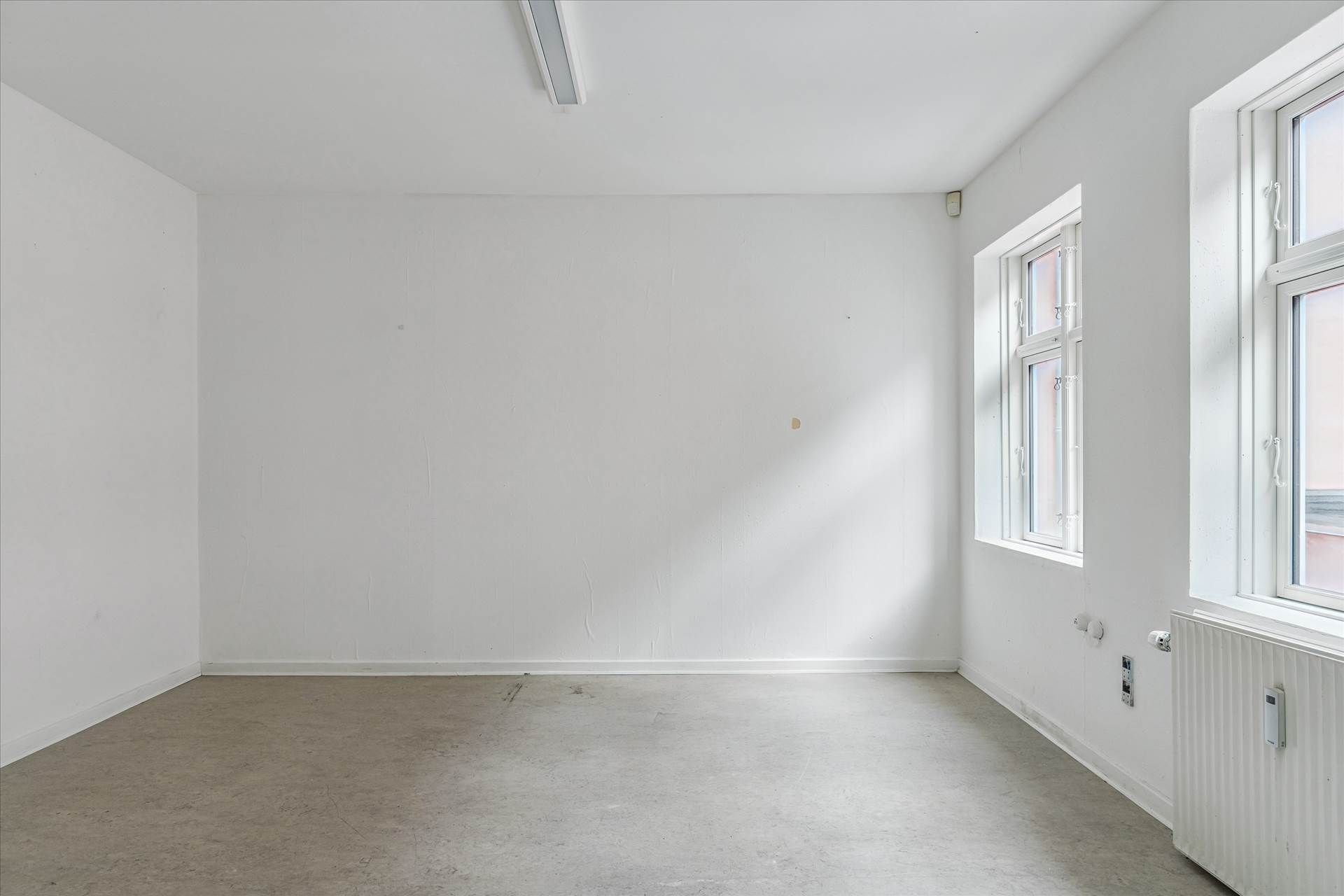 Hjørnerum med hvide vægge, gulv og loft, to vinduer, radiator og stikkontakter.