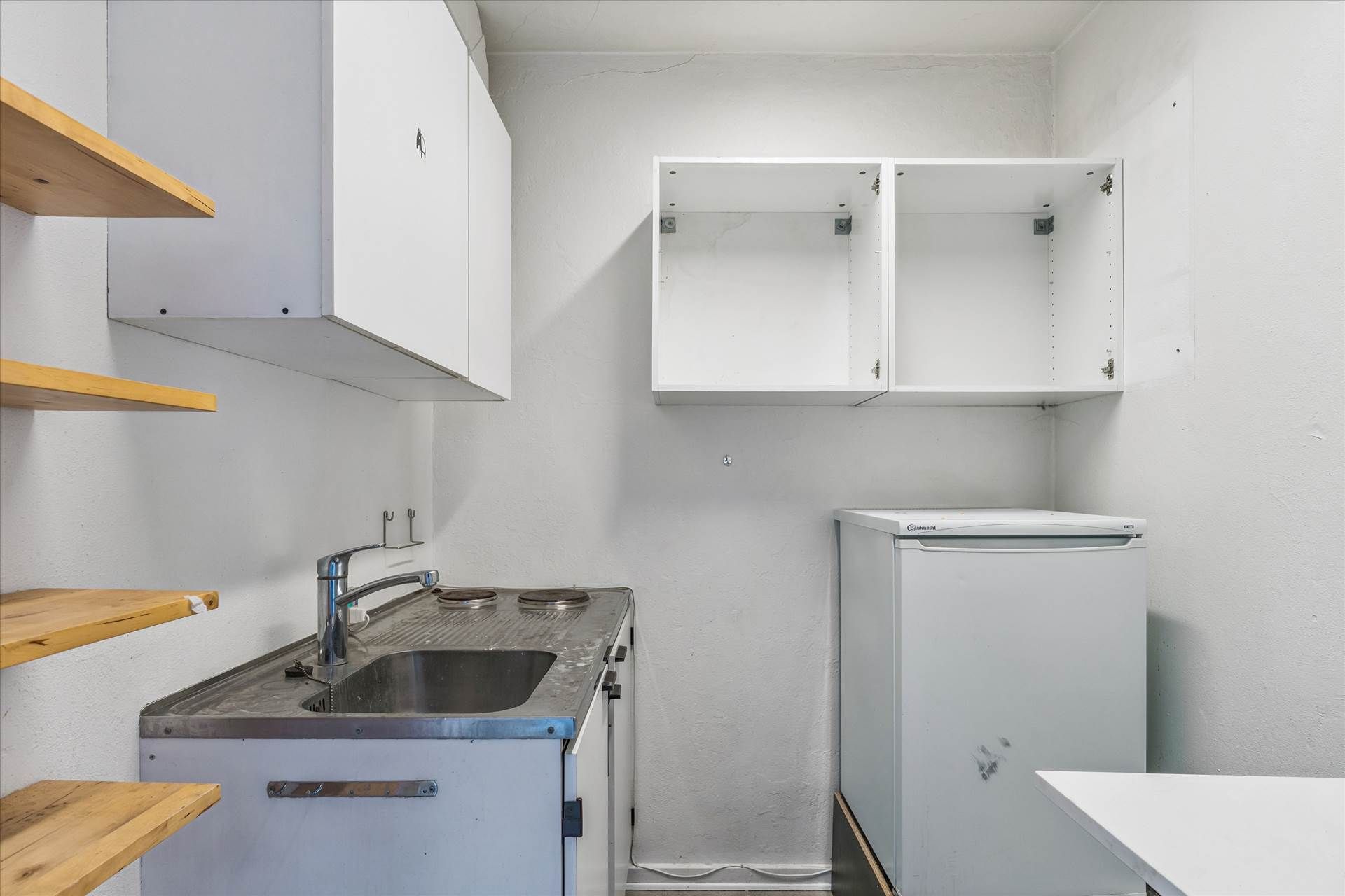 Et ubenyttet køkken med et rent gulv, en køleskab, en brændplads og tre højre træhylder