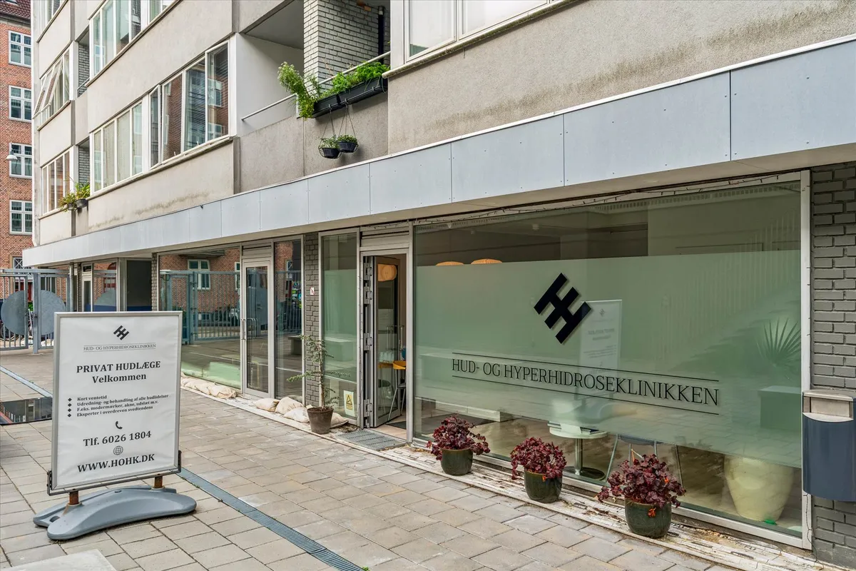 Privat hudlægeklinik med en indgang med glasdør og en skjoldformet logo samt fire lyserøde planter i potter foran vinduet