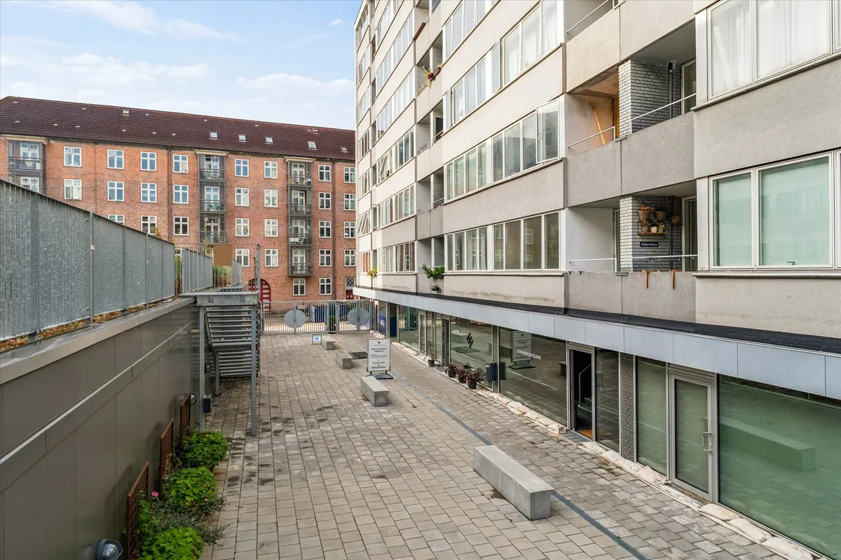 Bygning med store ruder og balkoner. Sted med brolægning, bænke og planter. En gård med to bygninger og trætrapper.