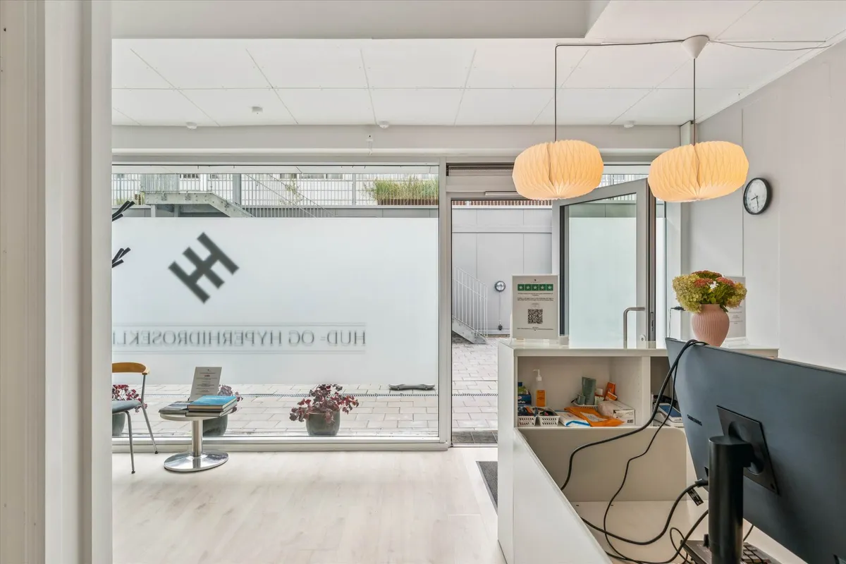 Inde i kontoret står en bord med blomster og en stol og en QR-kode på væggen. Der er to hængelampe og en klokke på væggen.