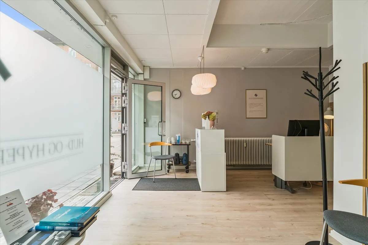 Indvendigt i et kontorlokal, der har et hvidt gulv, en åben dør og en skrivebord med en computer og en vase med blomster