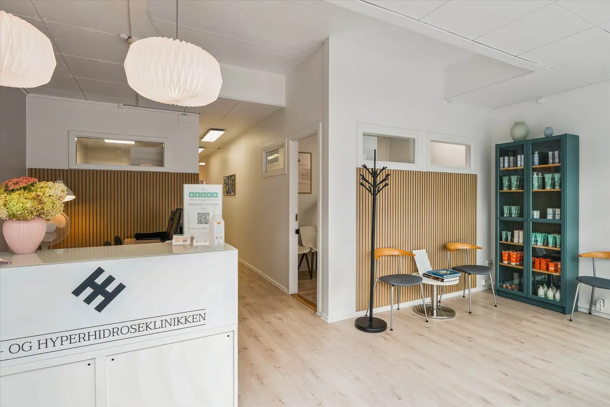 Indkørselsområde til et lægehus med hvide vægge og træpaneler, lysekroner og en skrivebord med et rødt H, en grøn skænk med