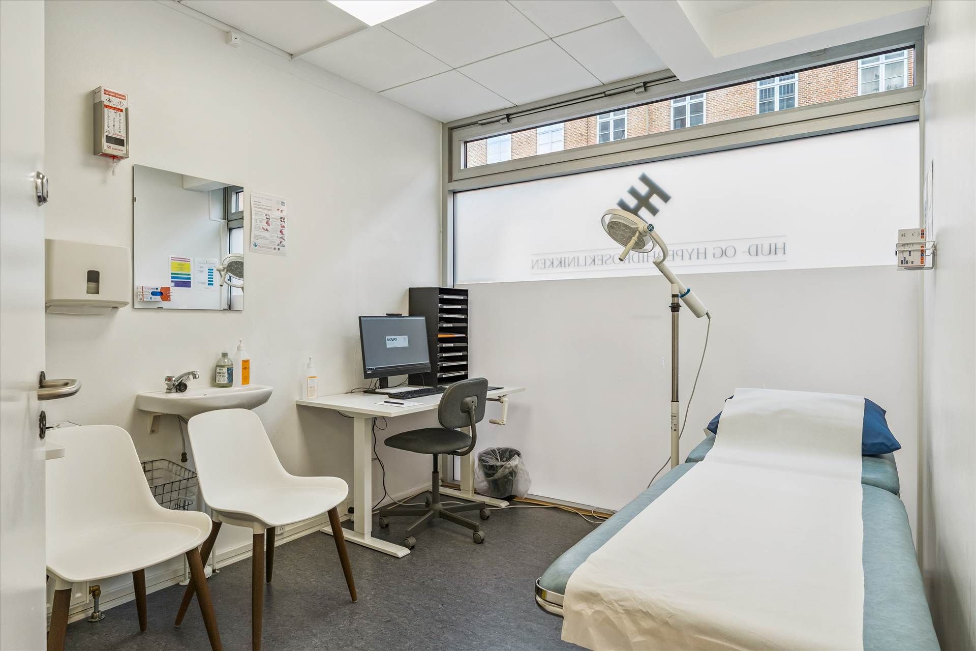 Hvidt lægeværelse med to stole, computerbord, strygejern, strygebræt, strygejernsstativ, strygebrætsholder, hængetoilet,