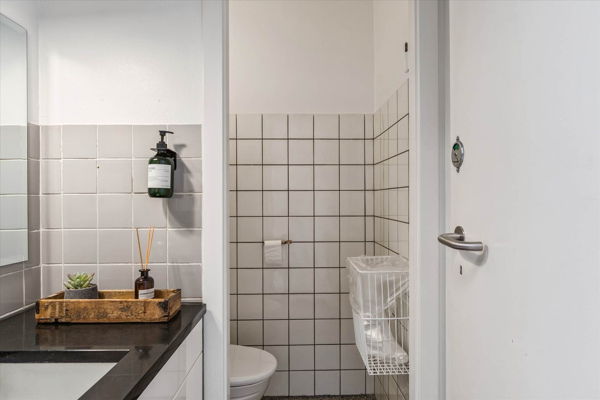 Et lille badeværelse med hvide flisede vægge, toalett, hængende håndsæbe og en lille plante i en gryde på en træbakke med