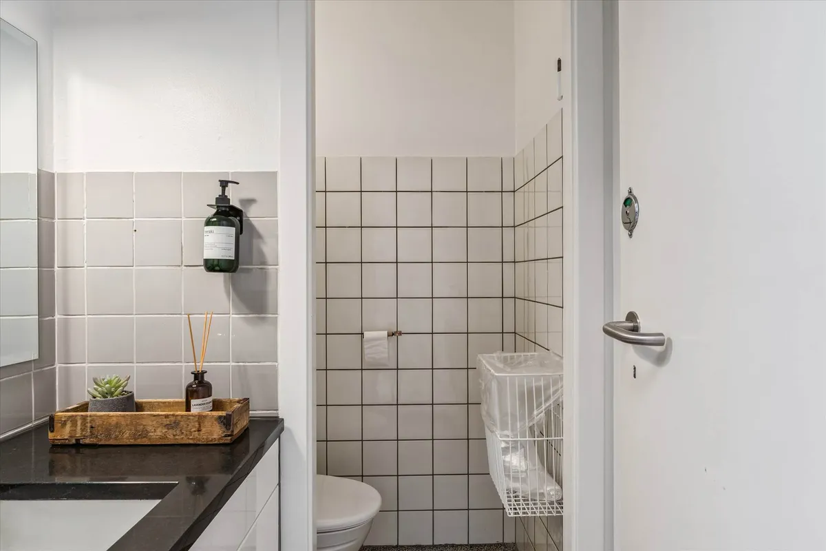 Et lille badeværelse med hvide flisede vægge, toalett, hængende håndsæbe og en lille plante i en gryde på en træbakke med