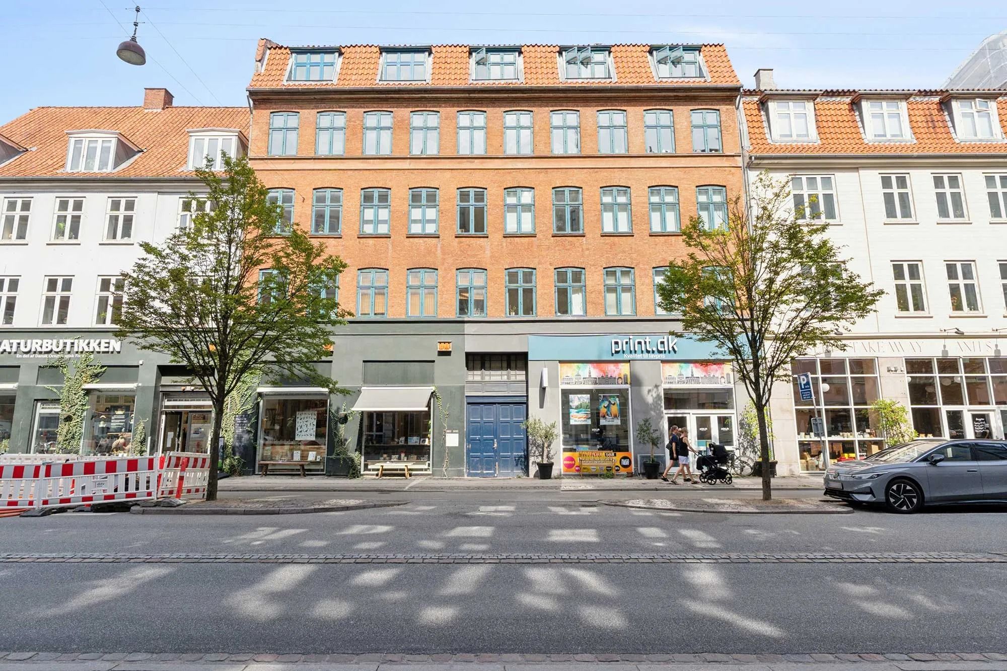 En husleje i København med grønne træer og forretninger langs gaden med biler og mennesker