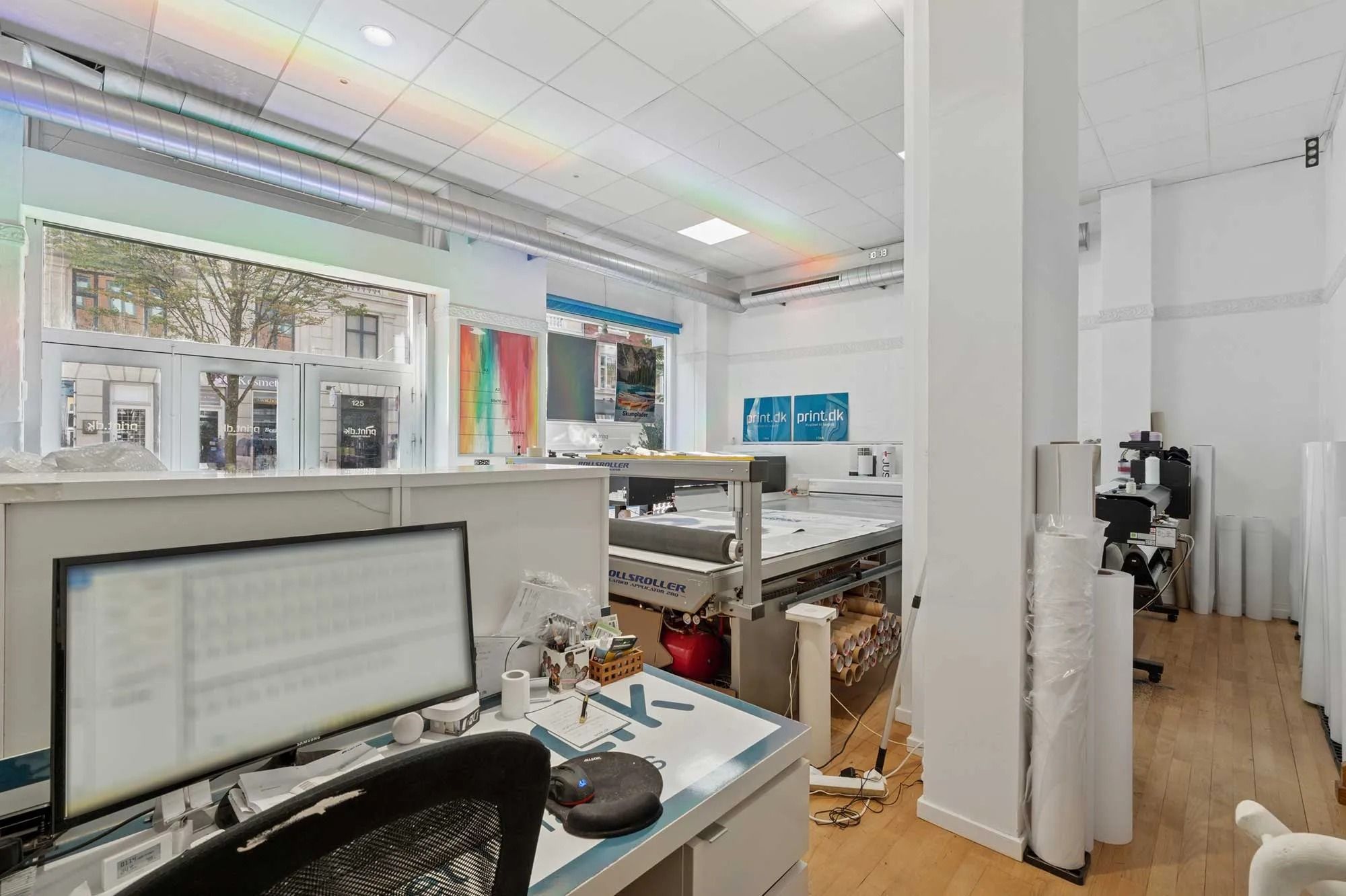 Et kontor med skrivebord, computer, stol, projektor og tavle. Plakater hænger på væggen, og der er kunstværker i baggrunden.