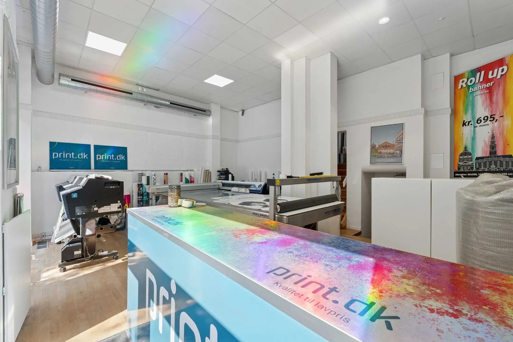 Københavns butik med print.dk-bannere på væggen, hvor en printer er placeret, og en regnbue er synlig på loftet