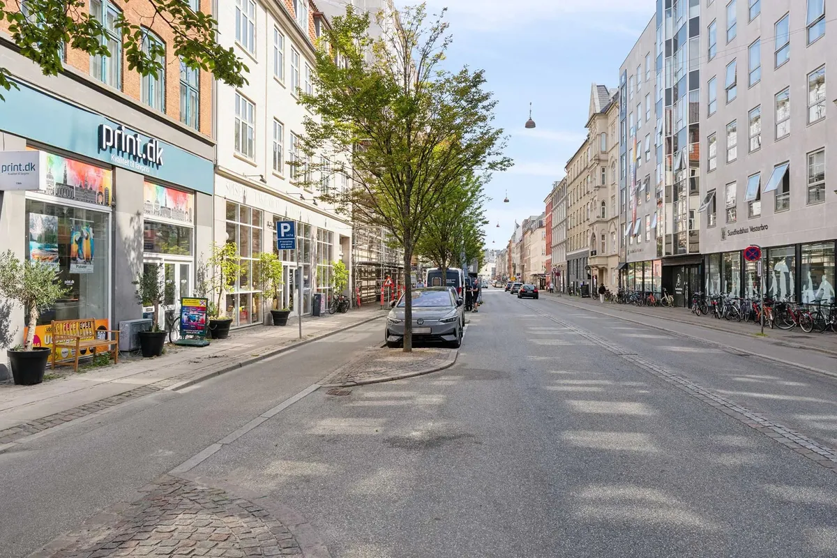 En tom gade med biler og cykler parkeret og forretninger langs gaden og grønne planter og træer på siderne