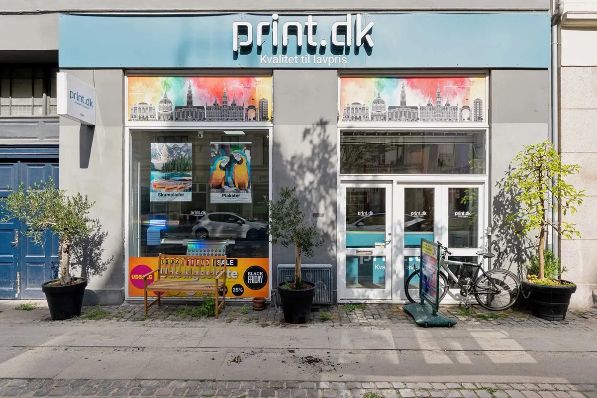 Butik med print.dk logo på facaden, med udsalgsskilt og rabatannoncering, og med to planter og en cykel foran dørene