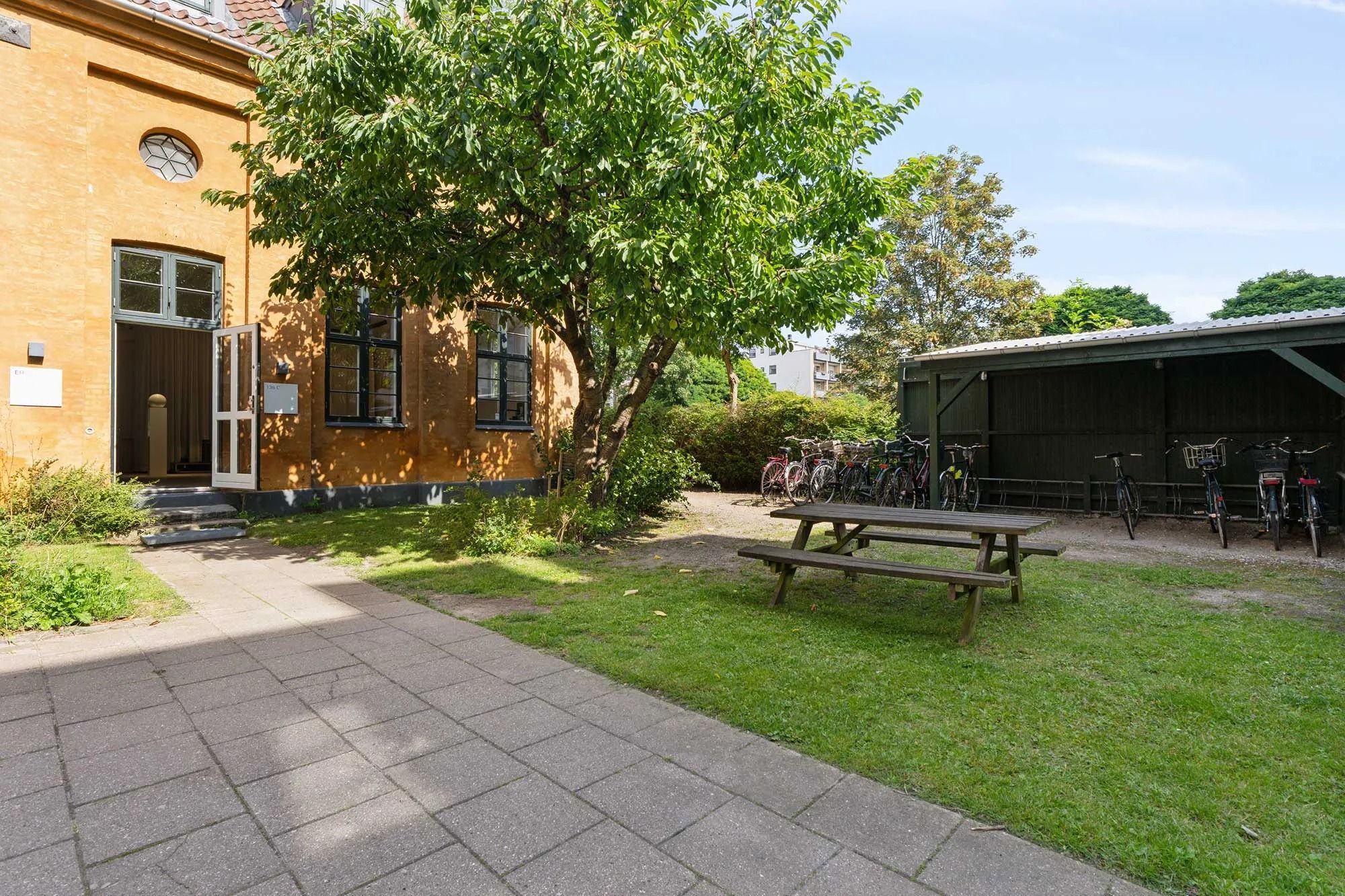 Lejlighed med grønne cykler og parkeringsplads foran træ og lysegul bygning med åben dør og vindue