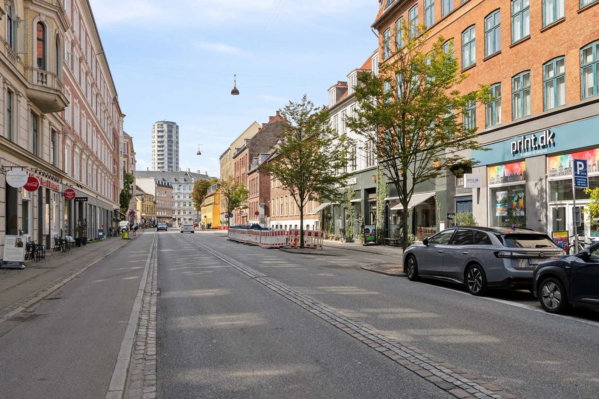 Strøget med forretninger og biler parkeret i København, med smukke træer og lyse himmel