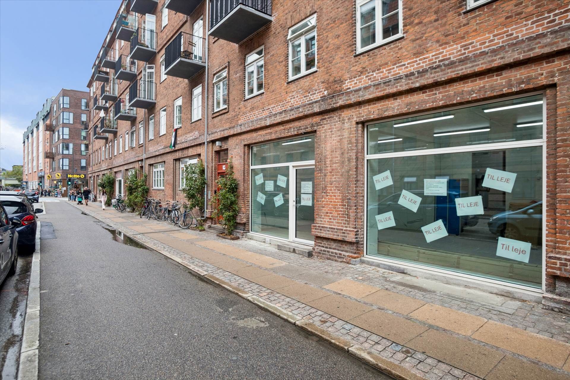 Butikslokaler med vinduer og skilte med teksten "Til leje" i et byområde med gadeblik og parkerede biler