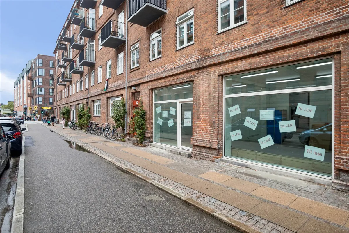 Butikslokaler med vinduer og skilte med teksten "Til leje" i et byområde med gadeblik og parkerede biler