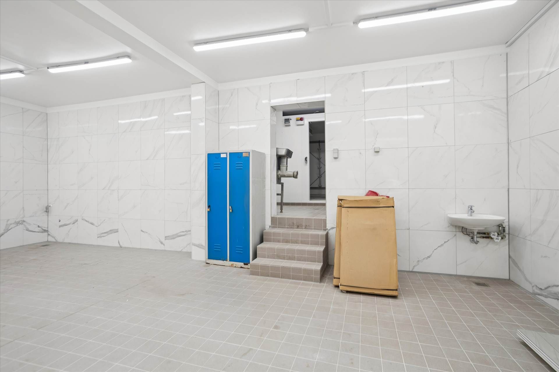 Rent og veludformet rum med hvide marmorvægge, hvidt loft, gulv med hvide firkantede fliser, lysekroner, stål skraldespand