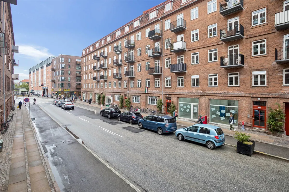 Adgangsvej med biler og cyklister, stående parkeret langs siderne og gågade med grønne planter og folk