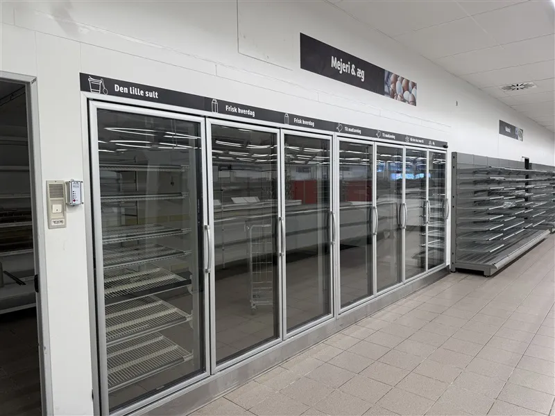 Køligt køleskab i dagligvarebutik med sort tekst "Den lille sult" og "Mejeri & æg" og krydsvise metalhylder og rulleskurve