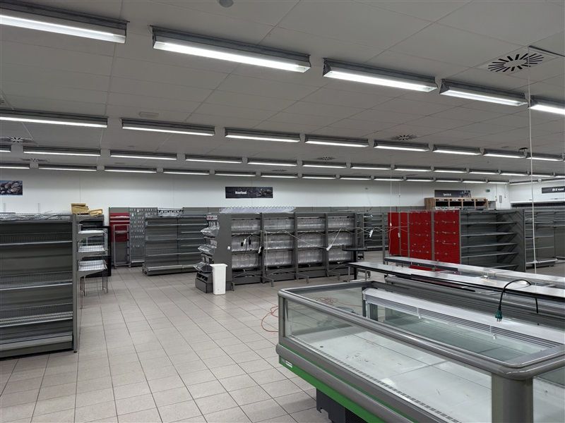Butikslokaler med tomme hylder og en stor fryseskab, belyst af lange, hvide lys. Gulvet er renset, og der er flere skilte på