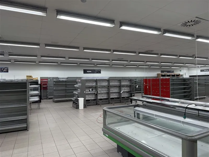 Butikslokaler med tomme hylder og en stor fryseskab, belyst af lange, hvide lys. Gulvet er renset, og der er flere skilte på