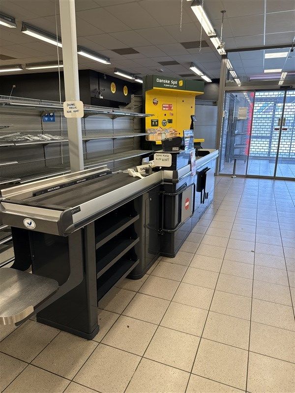 Butikslejemål med kassaskranke og varer til salg. Ingen kunder og tomme hylder. Forskelligt købmandsudstyr og gulvbelægning.