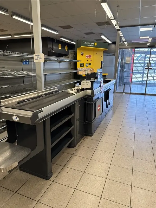 Butikslejemål med kassaskranke og varer til salg. Ingen kunder og tomme hylder. Forskelligt købmandsudstyr og gulvbelægning.