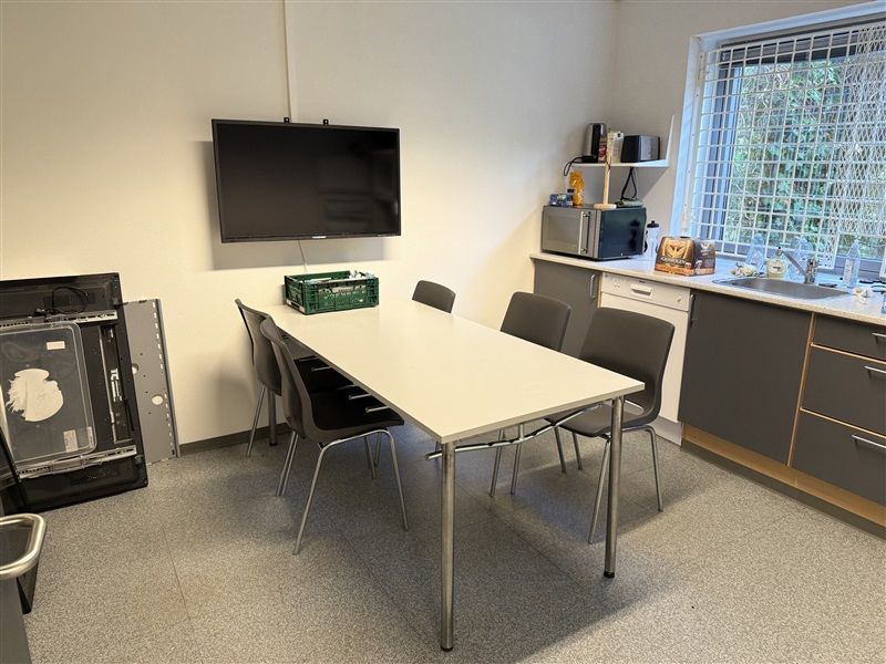 En moderne og veludstyret køkken med et stort hvidt bord, fire lyse grå stole og en stor tv-skærm på væggen