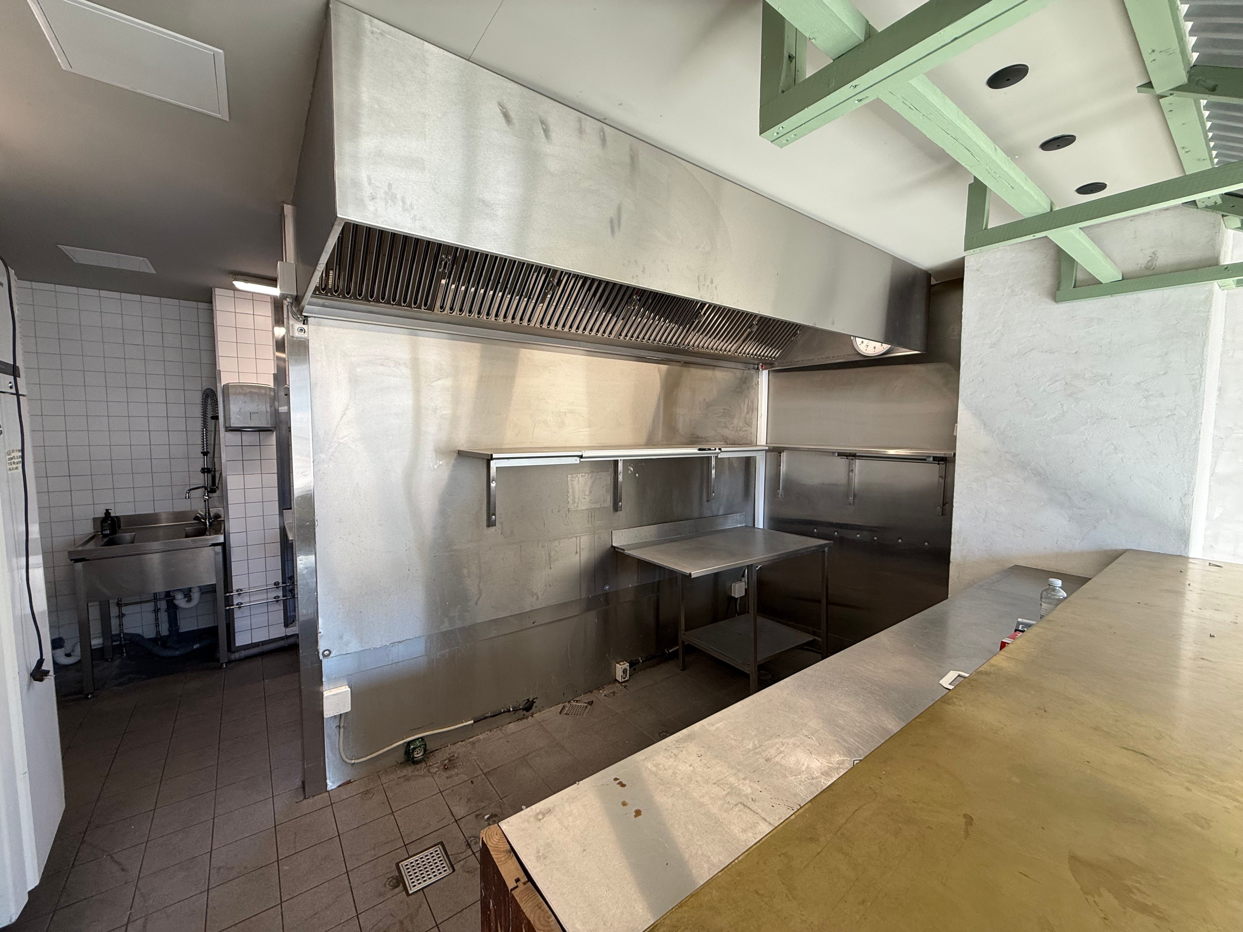 Take-away køkken med røgudsugning og arbejdsbord. Væggene er klinkelagde. Der er en stålvaskedam og stolpe med opvaske.