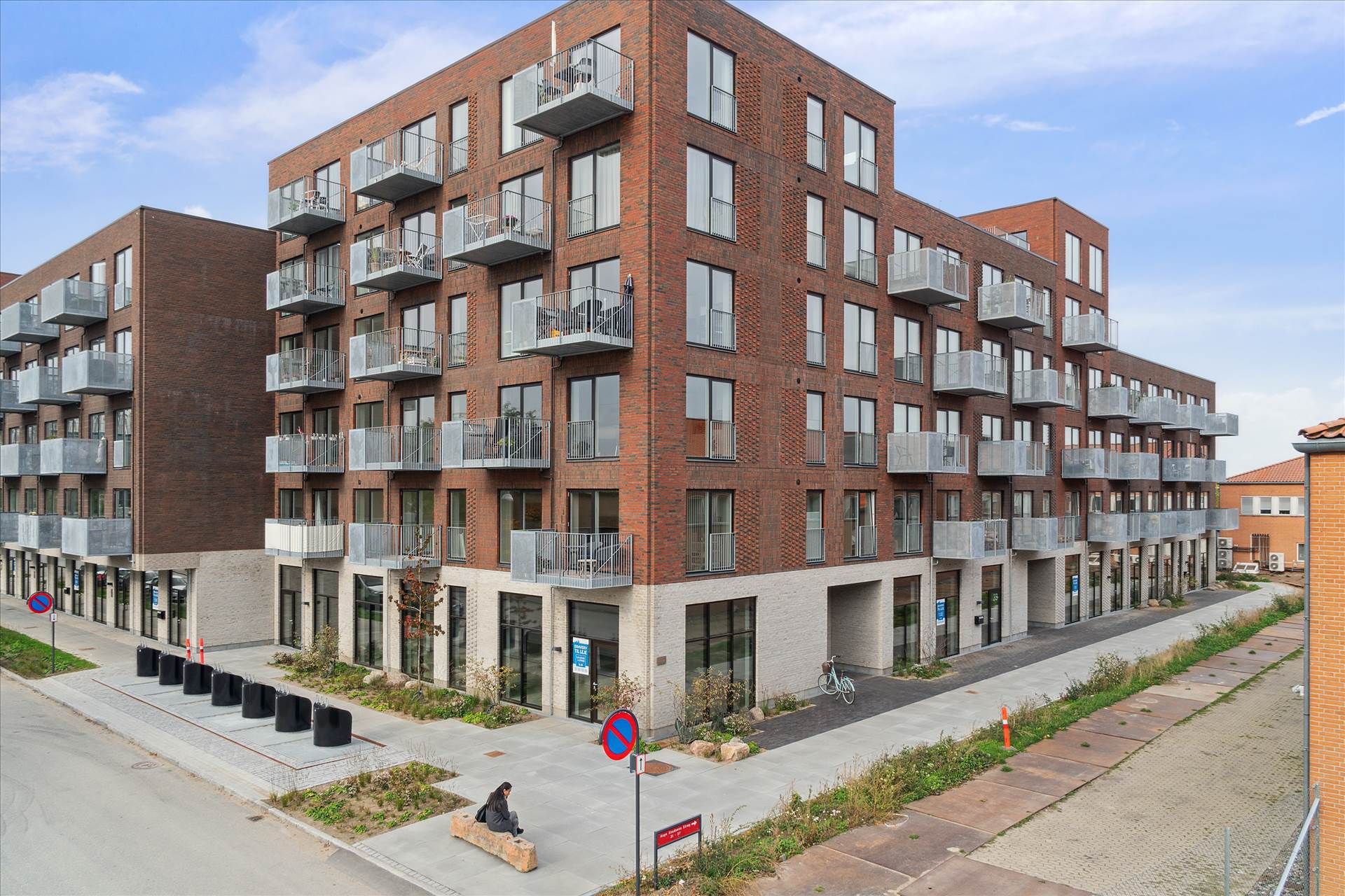 To høje bygninger med røde mursten og terrasser og glasvinduer. Person sidder på kantsten. På forgrunden står en cykel og
