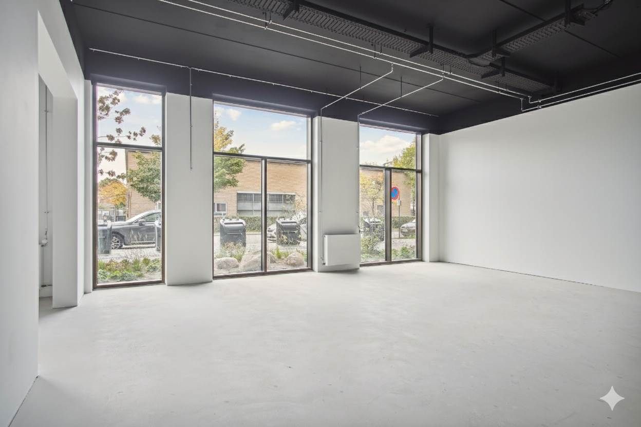 En tom lokale med hvide vægge, højt loft, glasvinduer og en stor indgang. Et skraldespand og parkerede biler er synlige