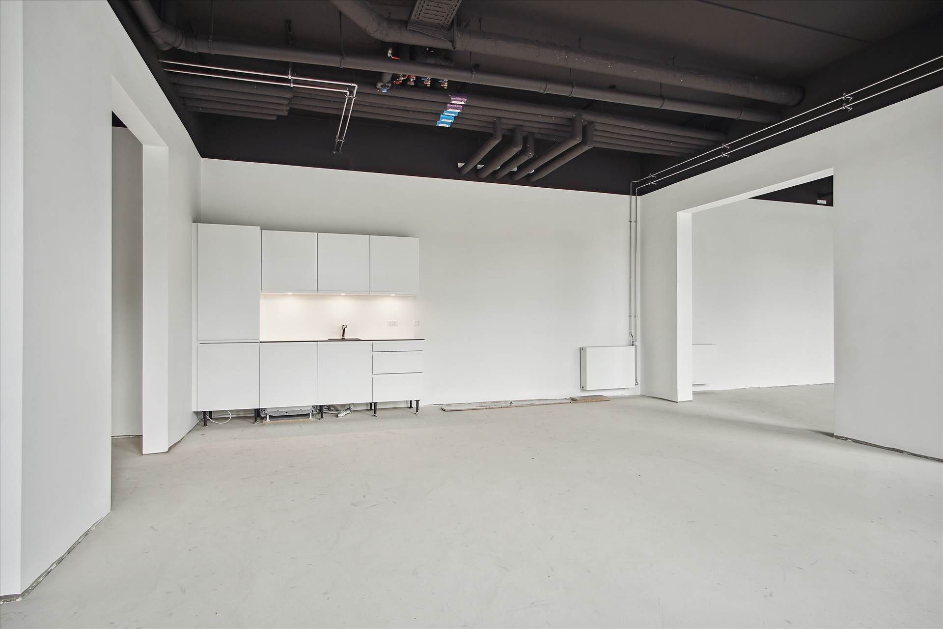 En stor, tom, hvid og rummelig værkstedslokal med hvide vægge og loft samt en hvid køkkenhjørne og et hvidt radiator.