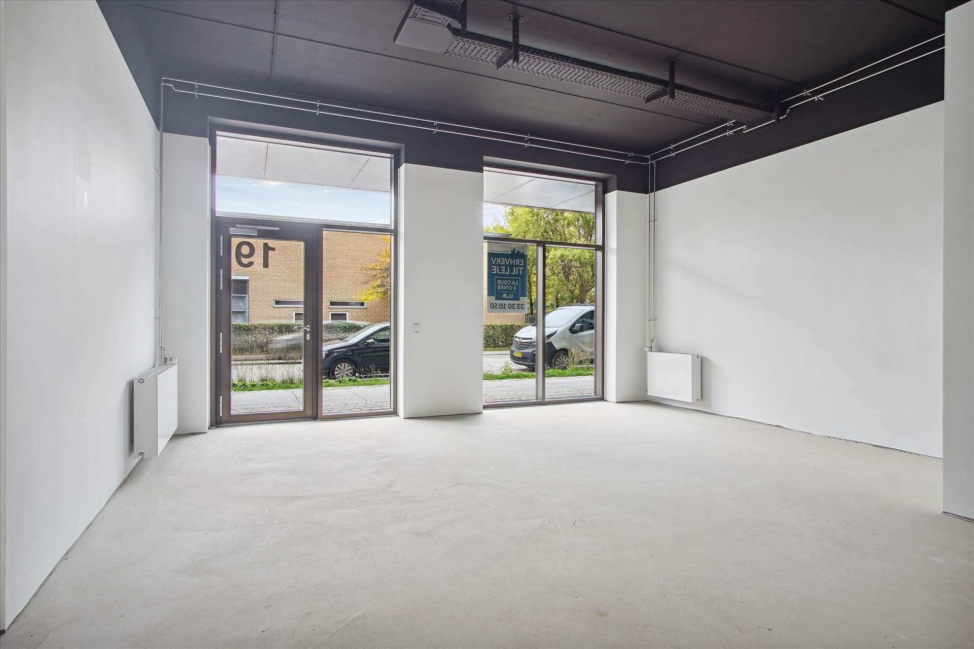 Tomme lokaler med glasdøre og vinduer, skilte på væggen, hvide vægge og møbleret med radiatorer og indretning.