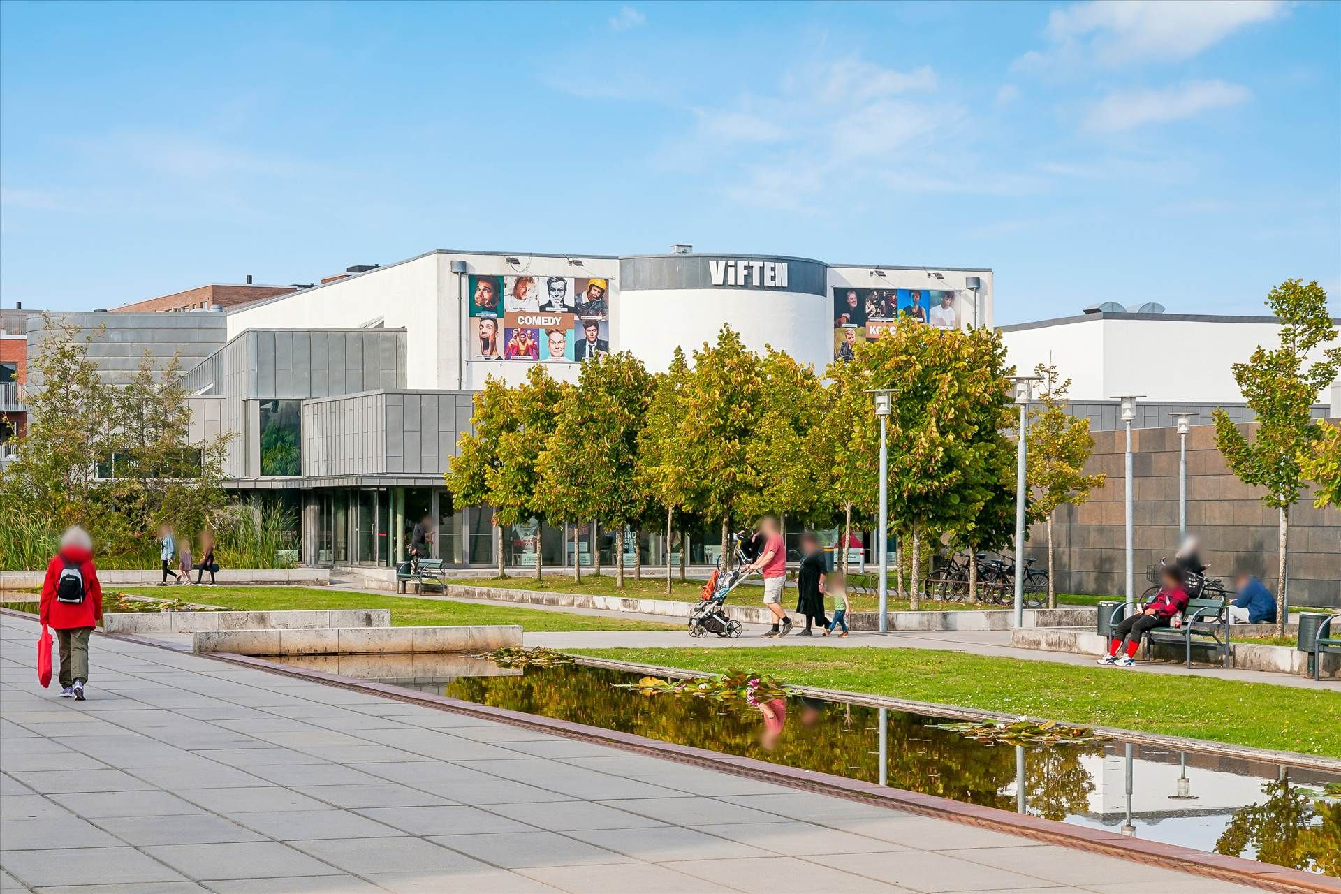 Familier og personer går forbi et komediehus med udstillinger på facaden, som hedder Viften. Parken foran er udstyret med