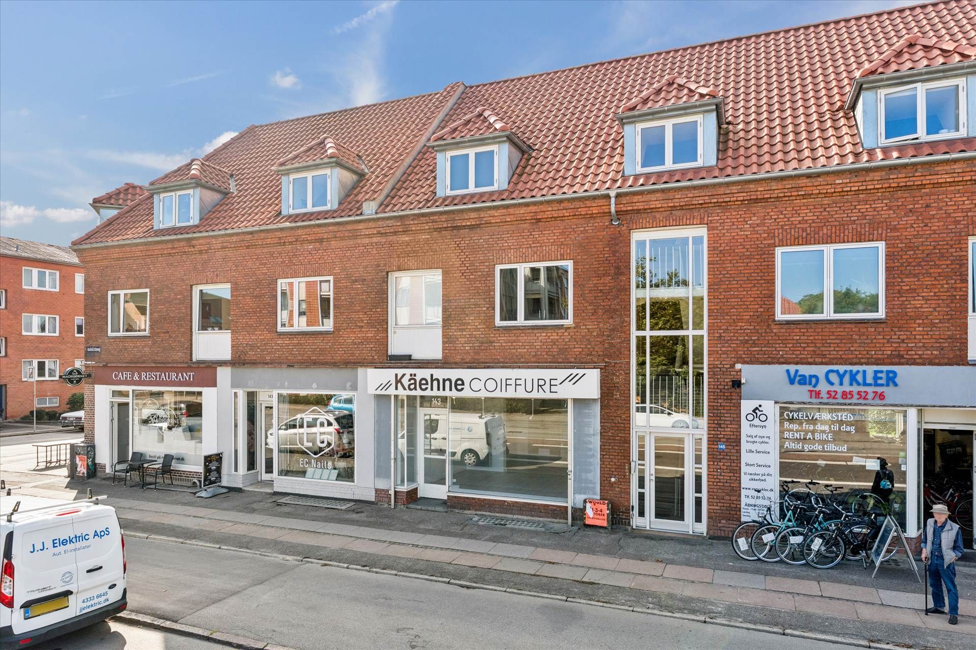 Et rødt teglbygget med to butikker og en kafé, hvor der står "Kæhne Coiffure" og "Van Cykler" på facaden. En mand står foran