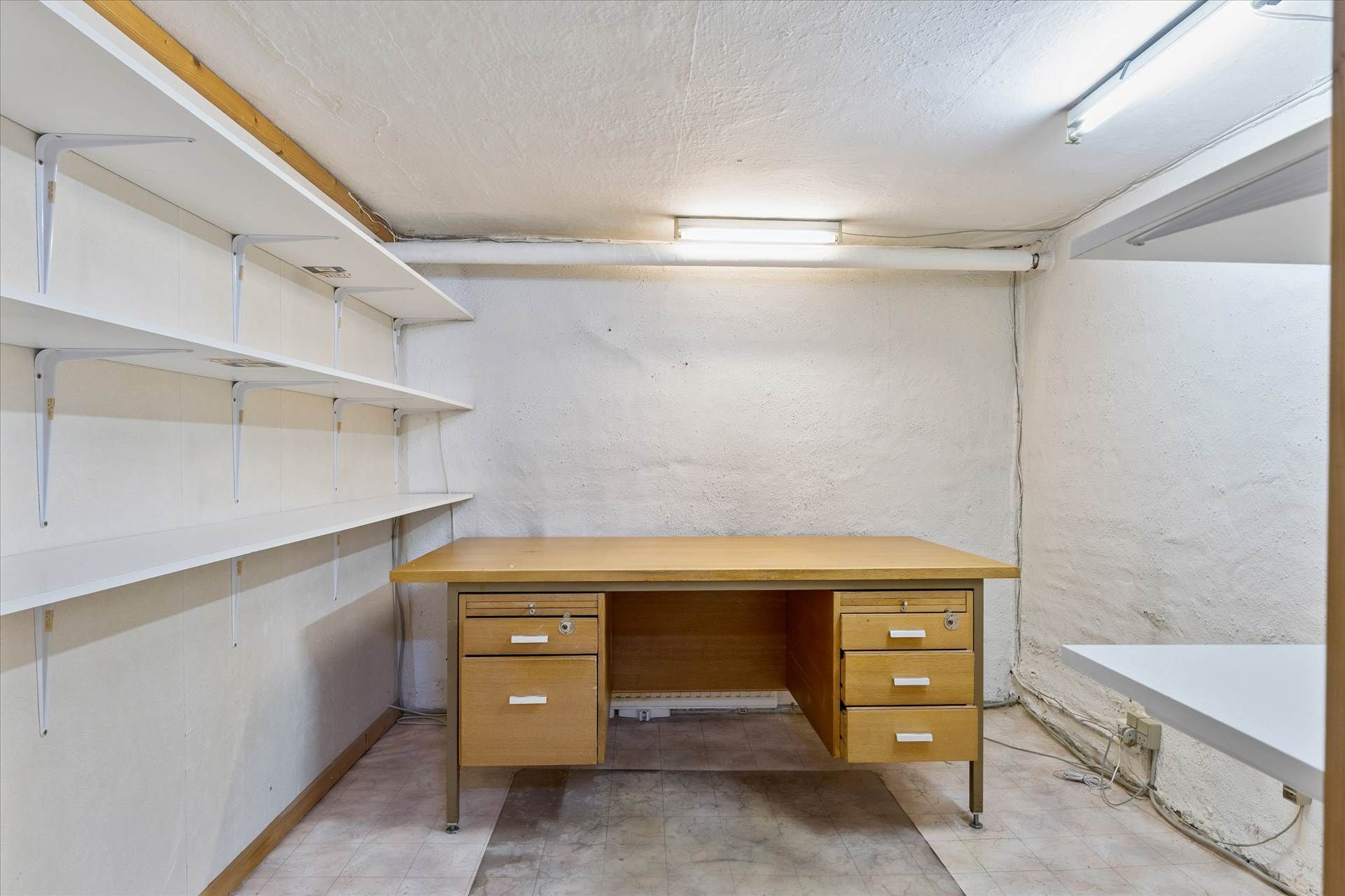 Lyst og rummeligt lokale med højt til loftet, hvidt loft og vægge, lyse lysekroner og trægulv. Et moderne skrivebord med