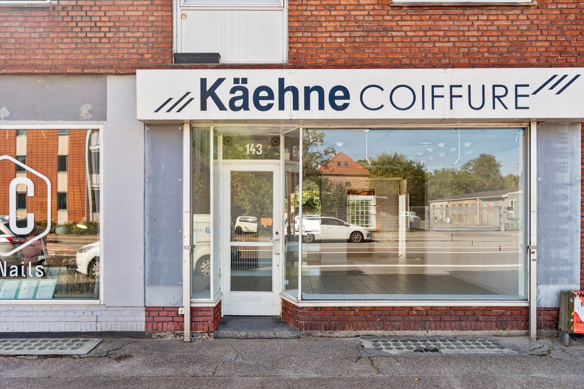 Forretningslokaler med skrift Kæhhne Coiffure på Ålekistevej, 143. Bygningen har glasvinduer og røde mursten. I spejlet ses