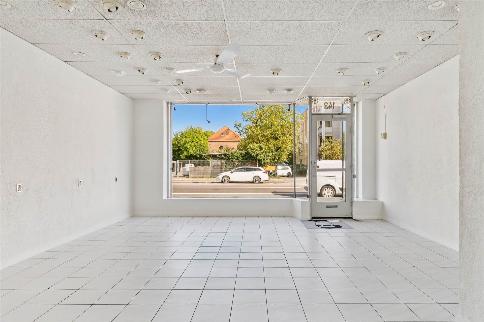 Lokale med hvidt loft, hvide vægge, og gulv. Dør i glasset og et vindue med udsigt til en bygning, en bil, og et træ